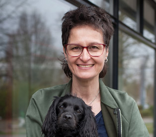 Rabea Wienholt, die einen schwarzen Hund mit langen Ohren auf den Armen hält, draußen vor einem Fenster stehend, mit Bäumen im Hintergrund.